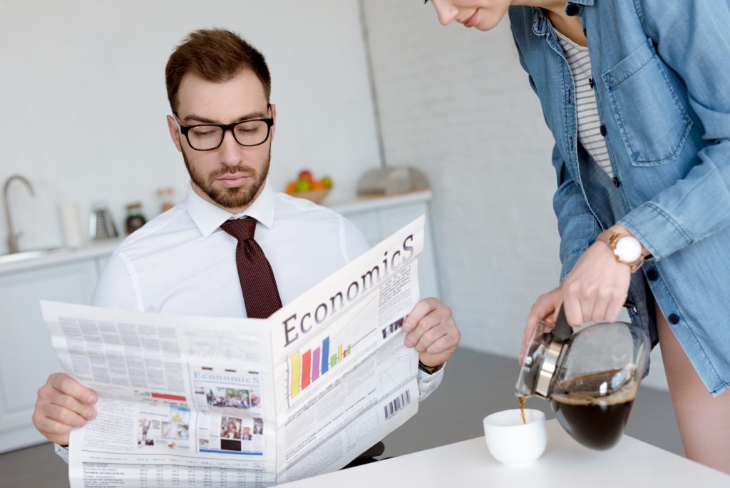 businessman in eyeglasses reading economics newspaper while wife pouring coffee in cup on kitchen