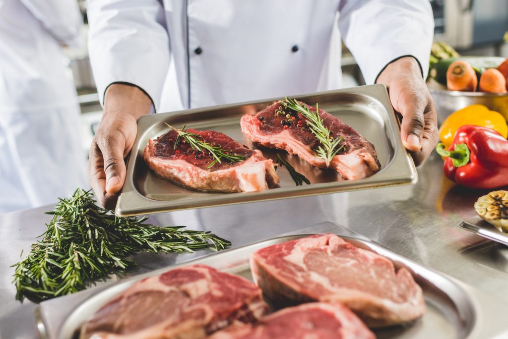 cropped image of african american chef holding tray with raw steaks at restaurant kitchen