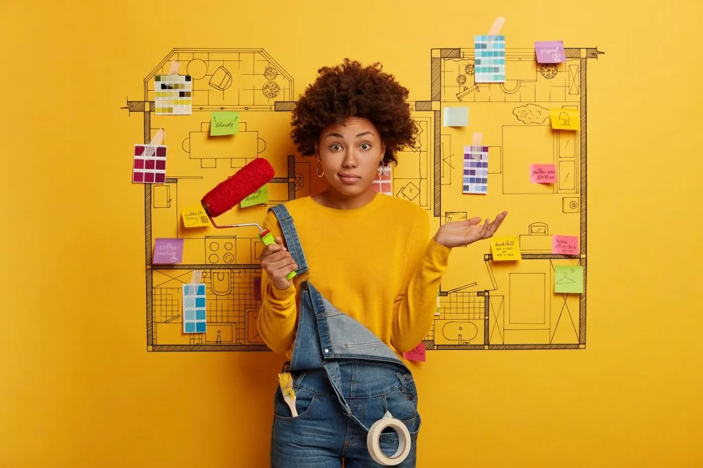 Hesitant curly woman stands with paint roller, thinks how to refurbish house, chooses new color for